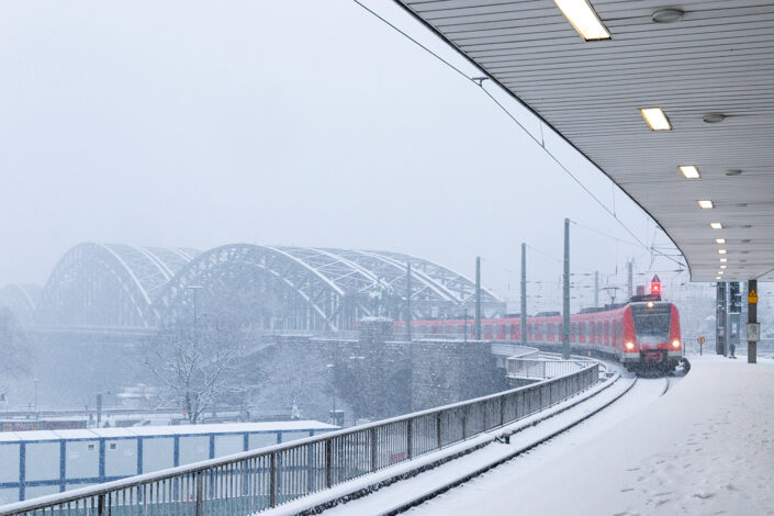 Einfahrender Zug an einem Gleis des Kölner Hauptbahnhofs. Im Hintergrund die Hohenzollernbrücke bei Schnee und Nebel