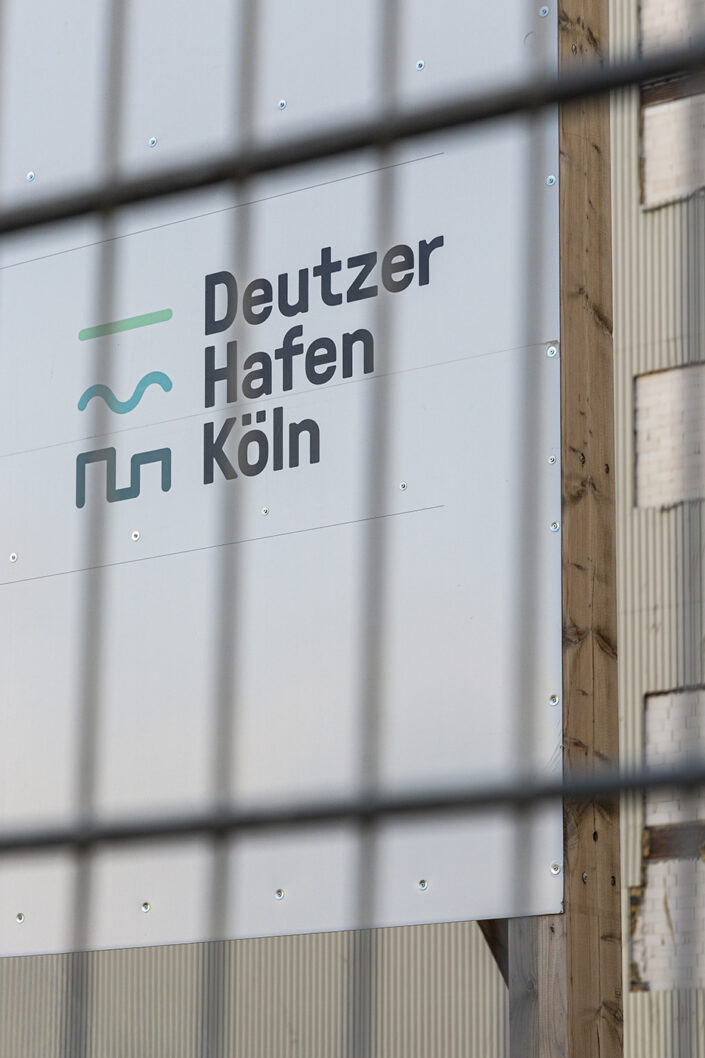 Banner mit der Aufschrift „Deutzer Hafen Köln“ hinter einem Bauzaun am Hafenareal.